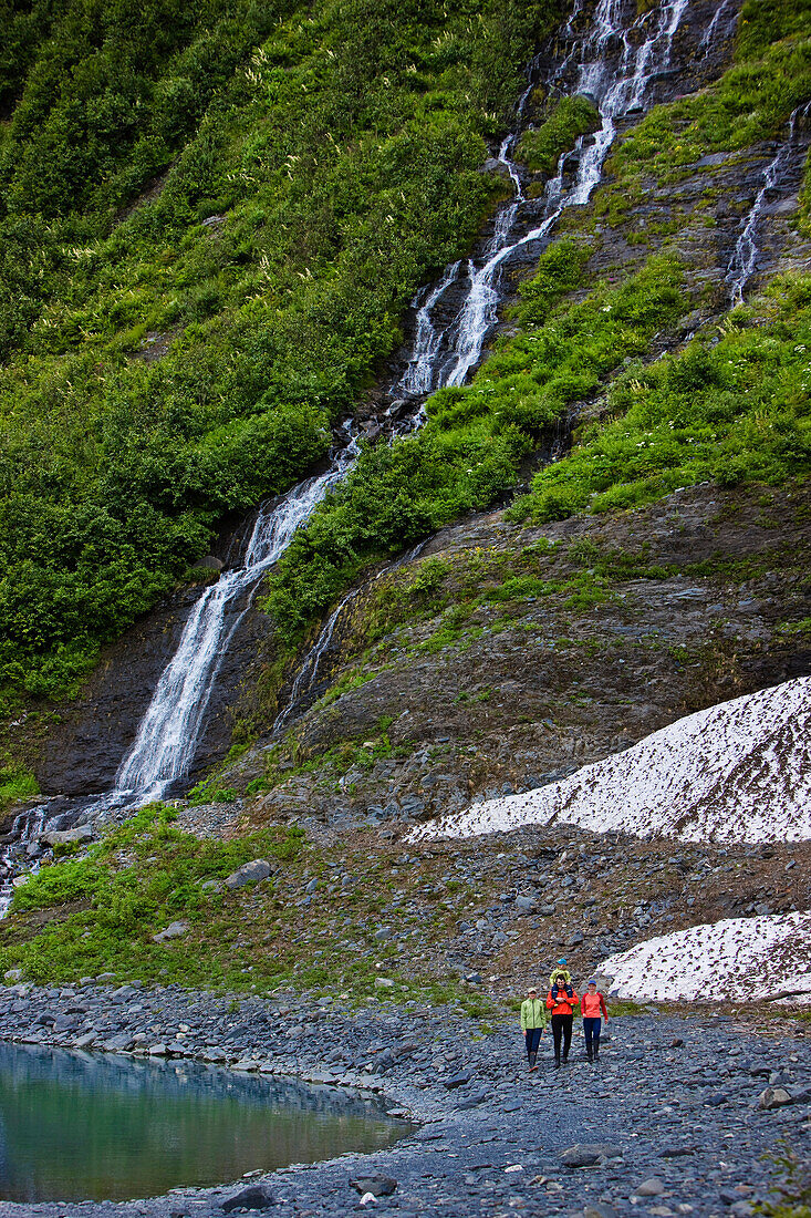 Group Of People Walking Along The Shoreline In Front Of A Waterfall, Shoup Bay State Marine Park, Prince William Sound, Alaska