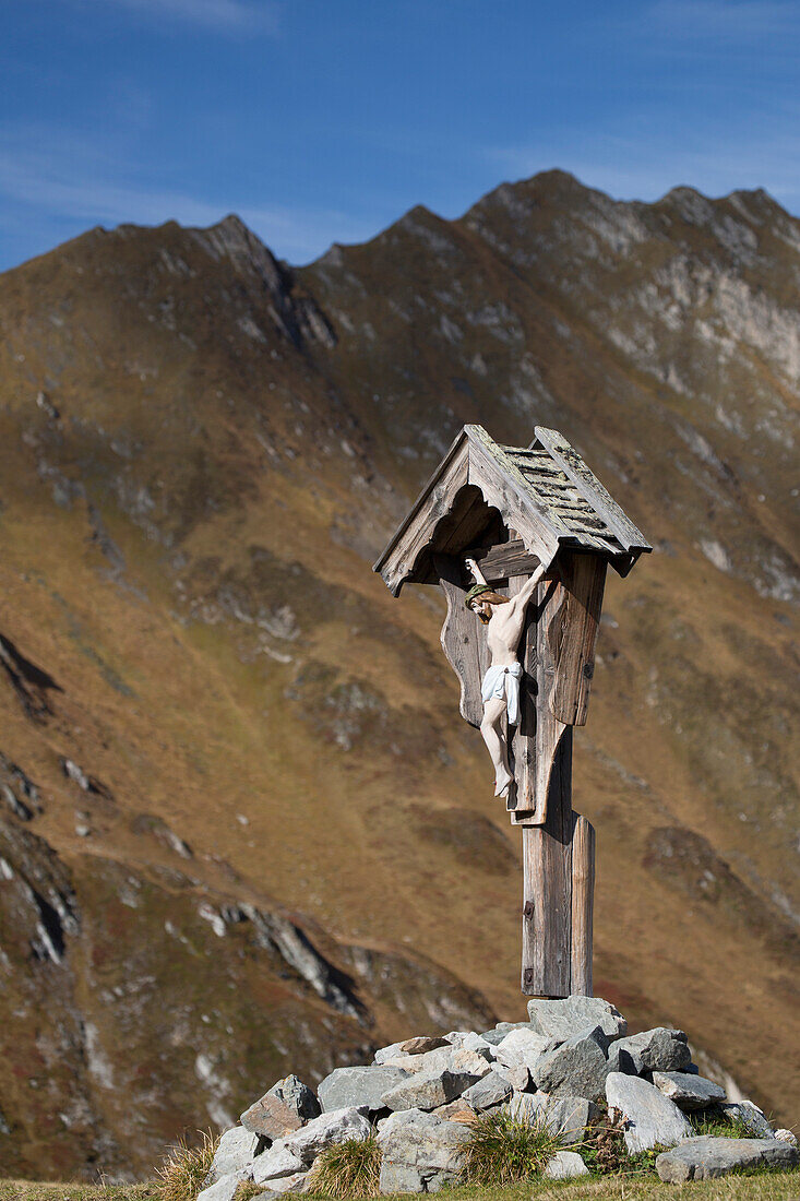 'Wooden crucifix memorial on grassy slope with mountain range and blue sky in the background; Hintertux, Austria'