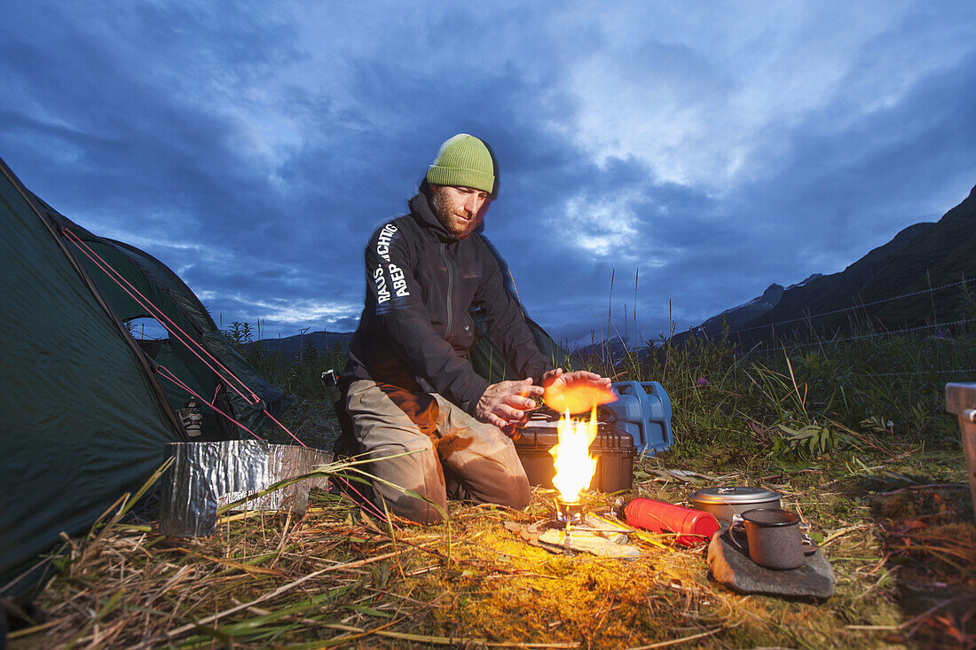 Swiss biologist and photographer warms hands while lighting camp stove along Kukak Bay at dusk, Katmai National Park, Southwest Alaska