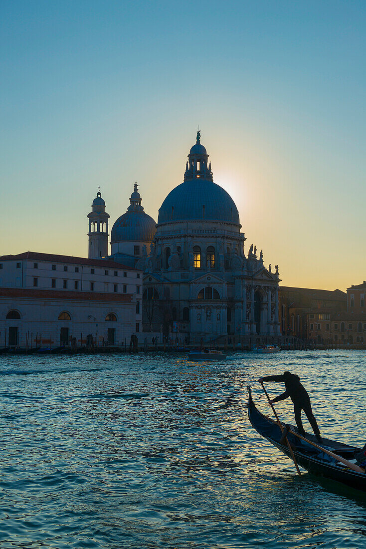 'Gondolier paddles his gondola with buildings and a church along the shoreline at sunset; Venice, Veneto, Italy'