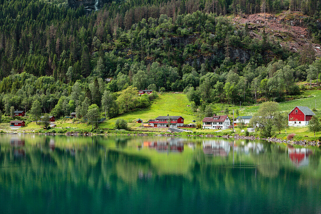 'The tranquil water of Nordfjorden reflecting the trees, houses and barn; Olden, Norway'