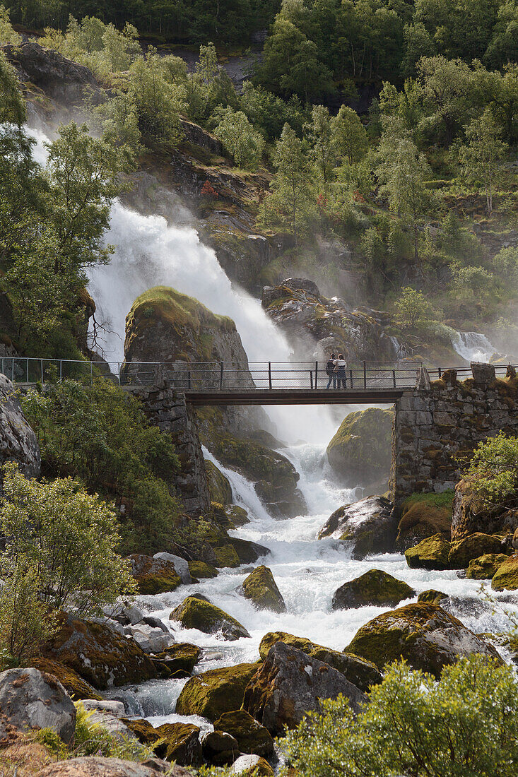 'Tourists stand on a bridge over a river in the mist of a waterfall; Olden, Norway'