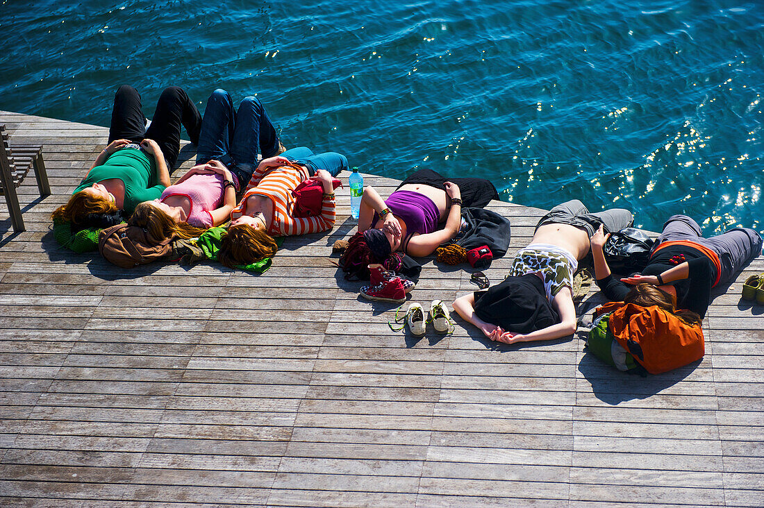 'A group of people laying on a wooden dock at the water's edge in the warm sunshine; Barcelona, Spain'