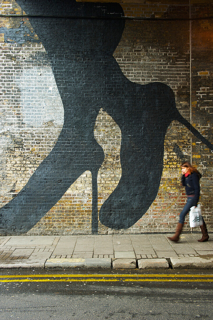 'A pedestrian walks by a brick wall with a woman's legs and feet in stilettos painted on the wall, Shoreditch; London, England'