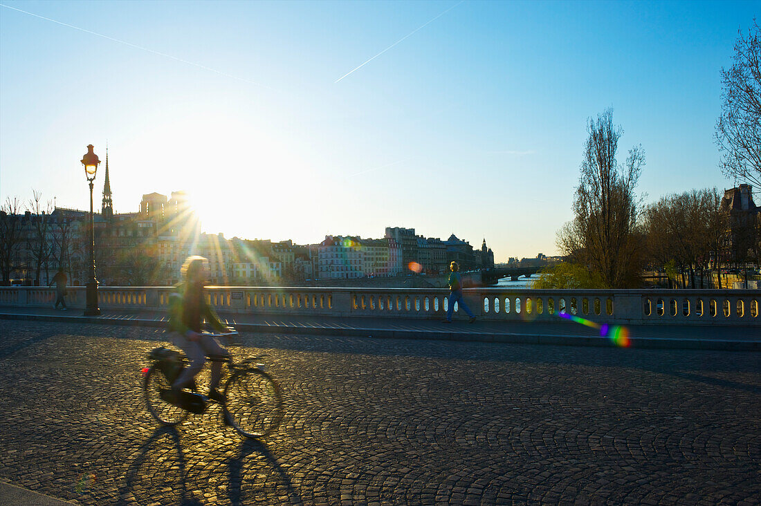 'A cyclist rides on a street across a canal at sunrise; Paris, France'