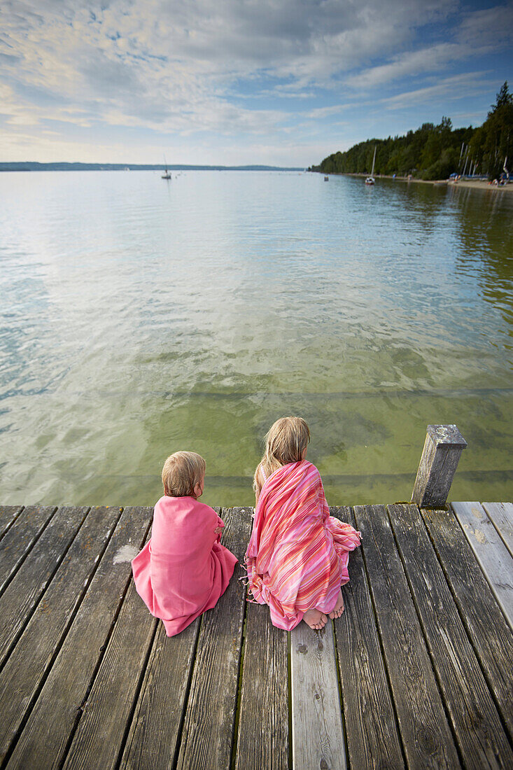 Two girls wrapped in towels sitting on a jetty at lake Starnberg, Upper Bavaria, Bavaria, Germany