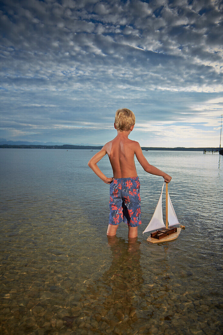 Boy with toy sail boat standing in lake Starnberg, Upper Bavaria, Bavaria, Germany