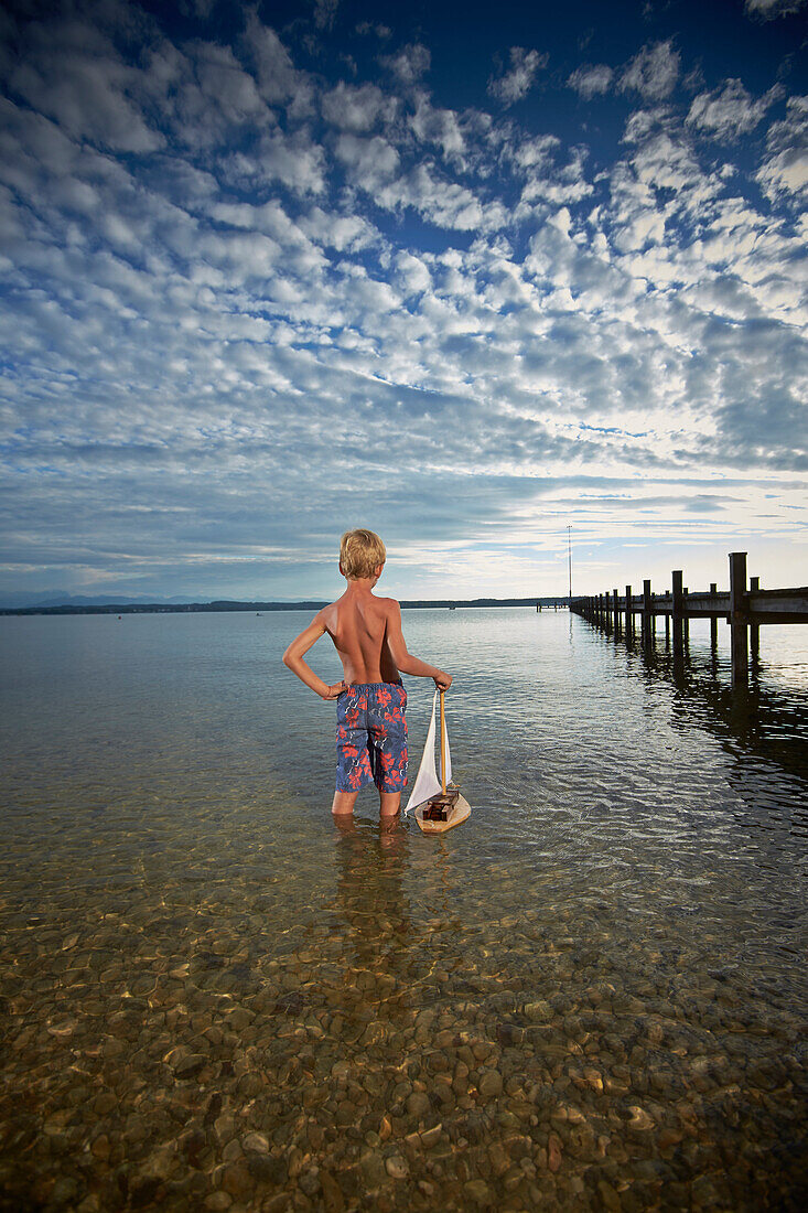 Boy with toy sail boat standing in lake Starnberg, Upper Bavaria, Bavaria, Germany