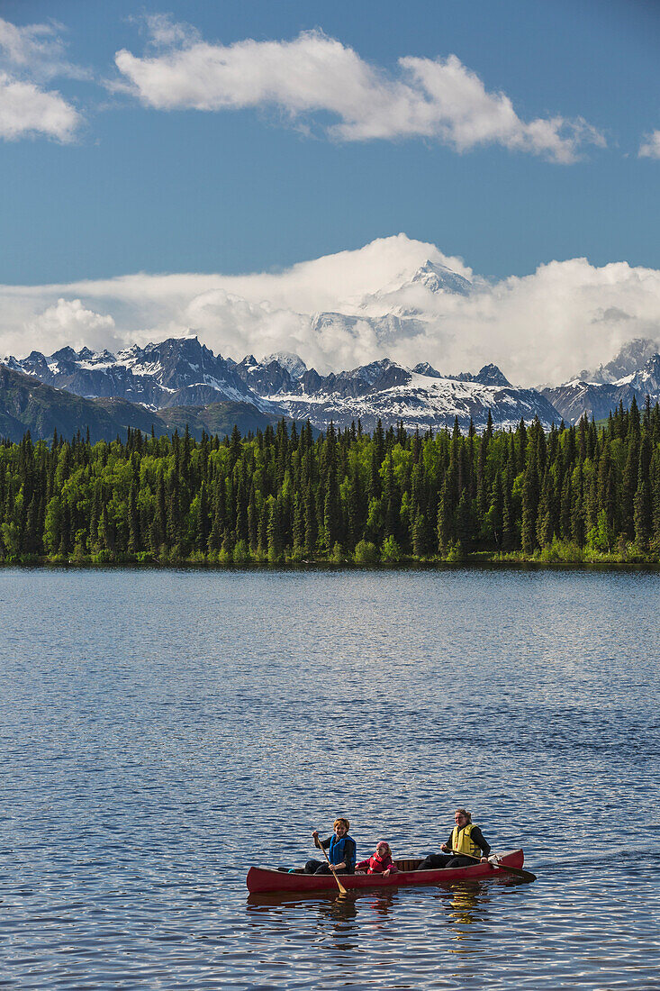 'Two women and young girl in a red canoe on Byers lake with green forested shoreline and Mount McKinley peaking through the clouds, Denali State Park; Alaska, United States of America'