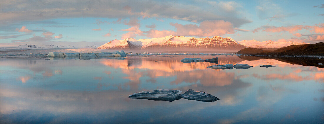 Panoramic view across the calm water of Jokulsarlon glacial lagoon towards snow-capped mountains and icebergs bathed in late afternoon light in winter, at the head of the Breidamerkurjokull Glacier on the edge of the Vatnajokull National Park, South Icela