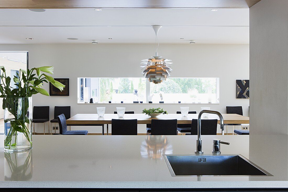 An open-plan kitchen - a sink in the kitchen counter and view of the dining table with a designer lamp