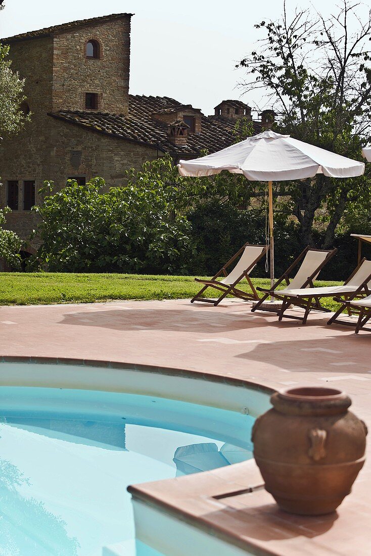Clay planter beside a pool and lounge chairs under a sun umbrella with a view of a Mediterranean grange
