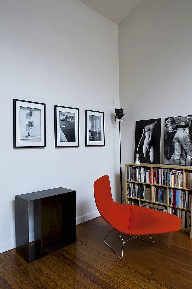 Corner of a living room with a lounger upholstered in red in front of half-high bookshelves and photos on the wall
