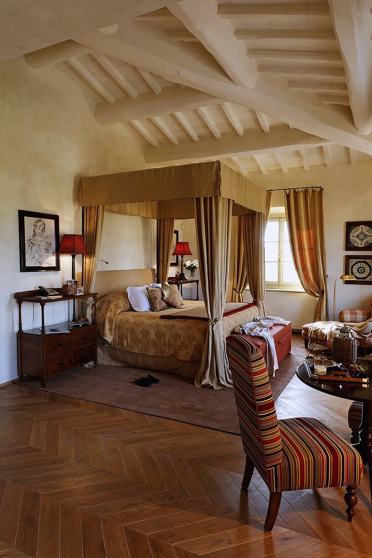 Canopy bed under a white beam ceiling and armchairs with a side table on a herring bone floor