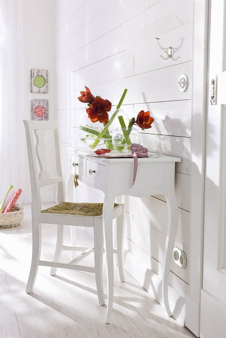 A white wall table and a chair against a wood panelled wall and red amaryllis in a glass vase