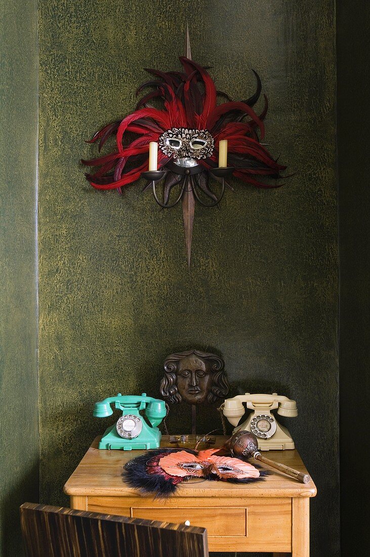 A wall table made of light wood with old telephones and a mask hanging from a candle holder on a dark wall