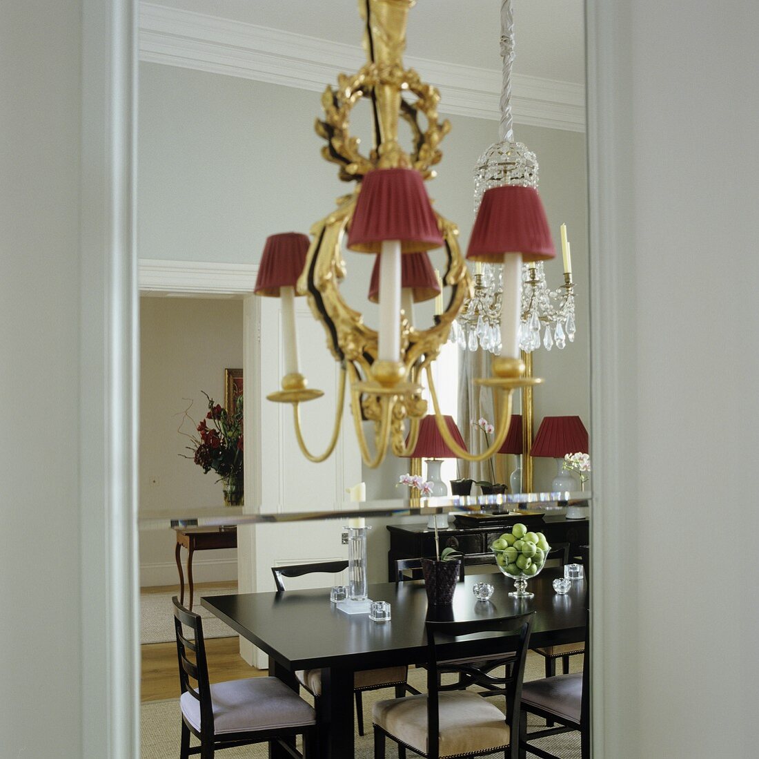 A brass chandelier with red shades mounted on a mirror in which a black dining and chairs are reflected