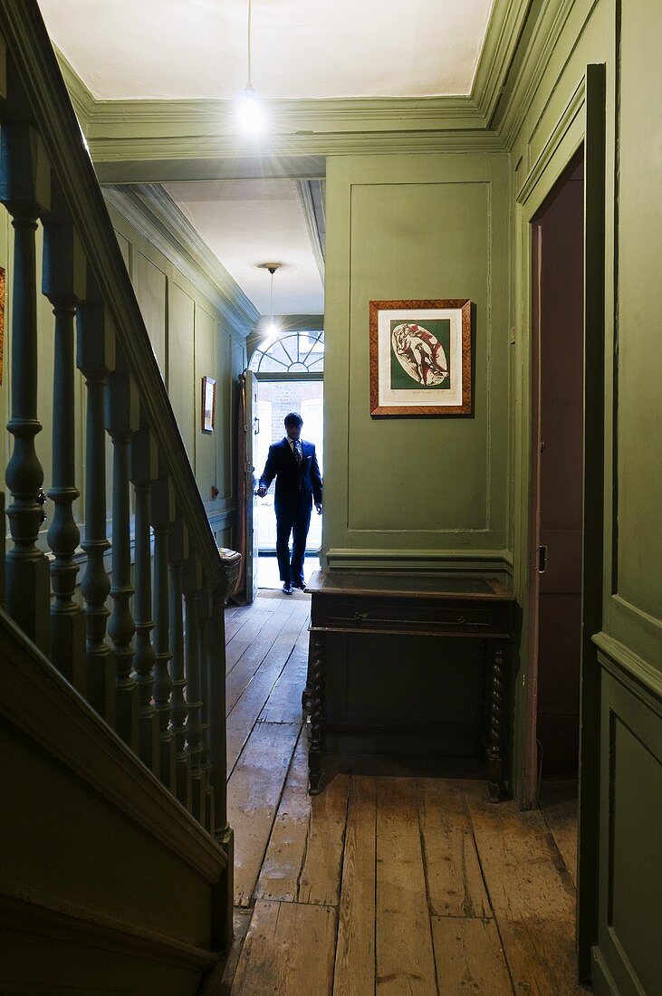 Green wood panelling in a corridor with a flight of old wooden stairs and a man at the front door