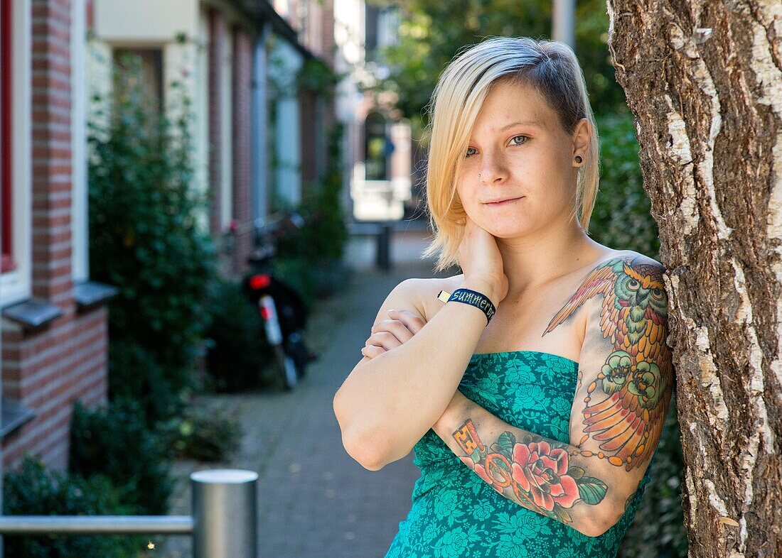 Tilburg, Netherlands. Young blonde woman with a tattoo on her left arm leaning against a tree in a domestic street.