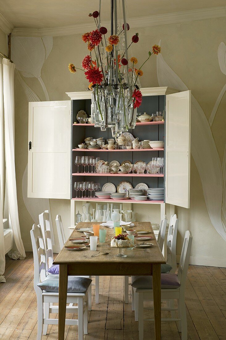 A wooden table and white chairs in front of a crockery cupboard with open doors