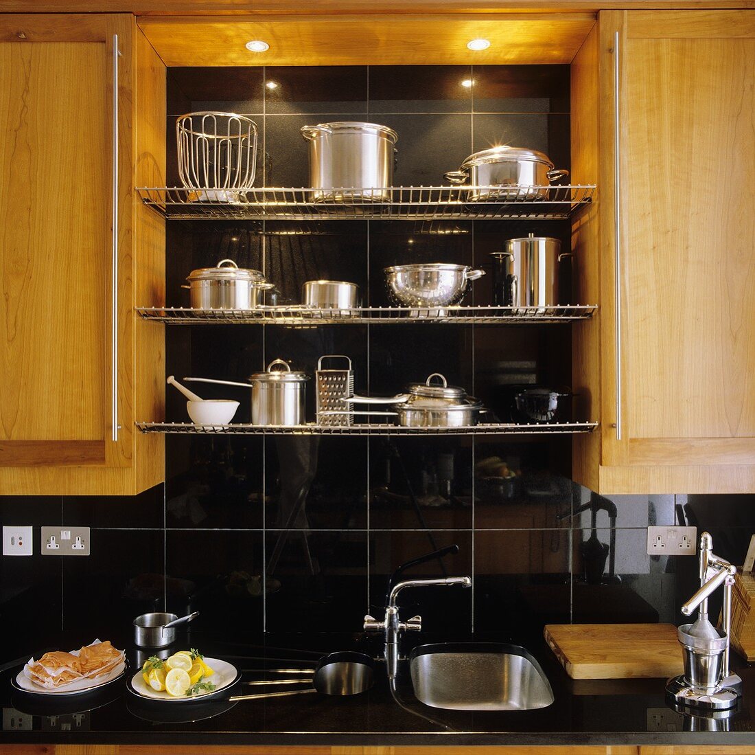 Kitchen cupboards with wooden doors with stainless steel pans on a wire shelf in a black-tiled niche