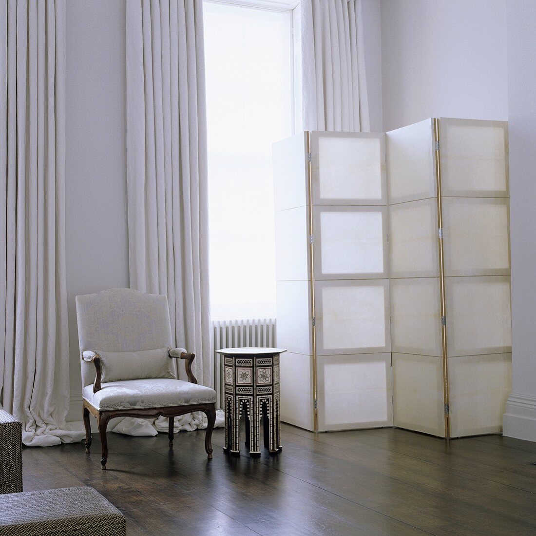 A corner of a living room with a white paravent and an antique armchair in front of a window