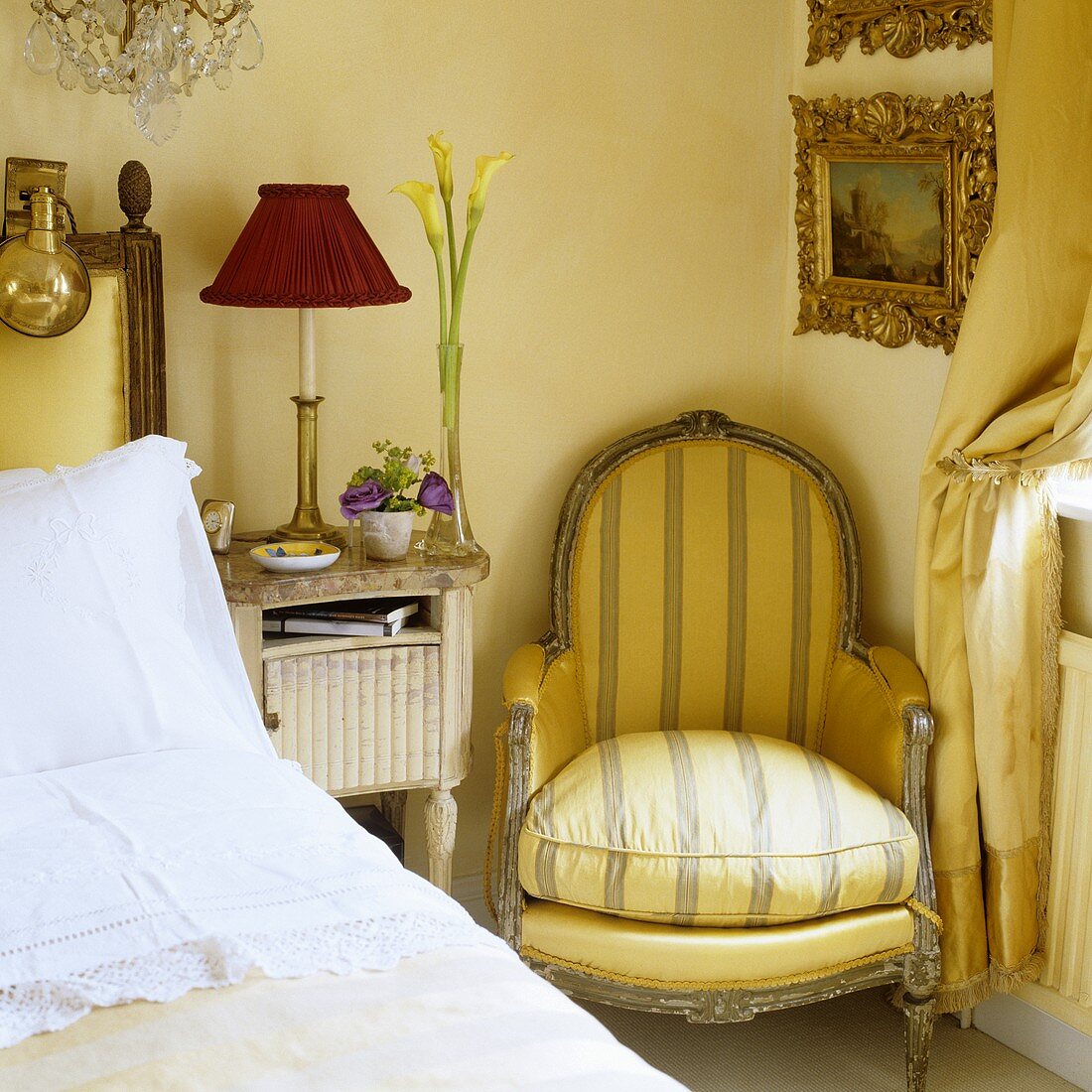 An antique armchair next to a bedside table with a lamp with a red shade in the corner of a bedroom