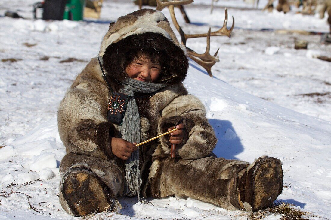 mit Rentierfellen bekleidetes Nomaden-Mädchen sitzt im Schnee, Tschukotka (Autonomer Kreis der Tschuktschen), Sibirien, Russland