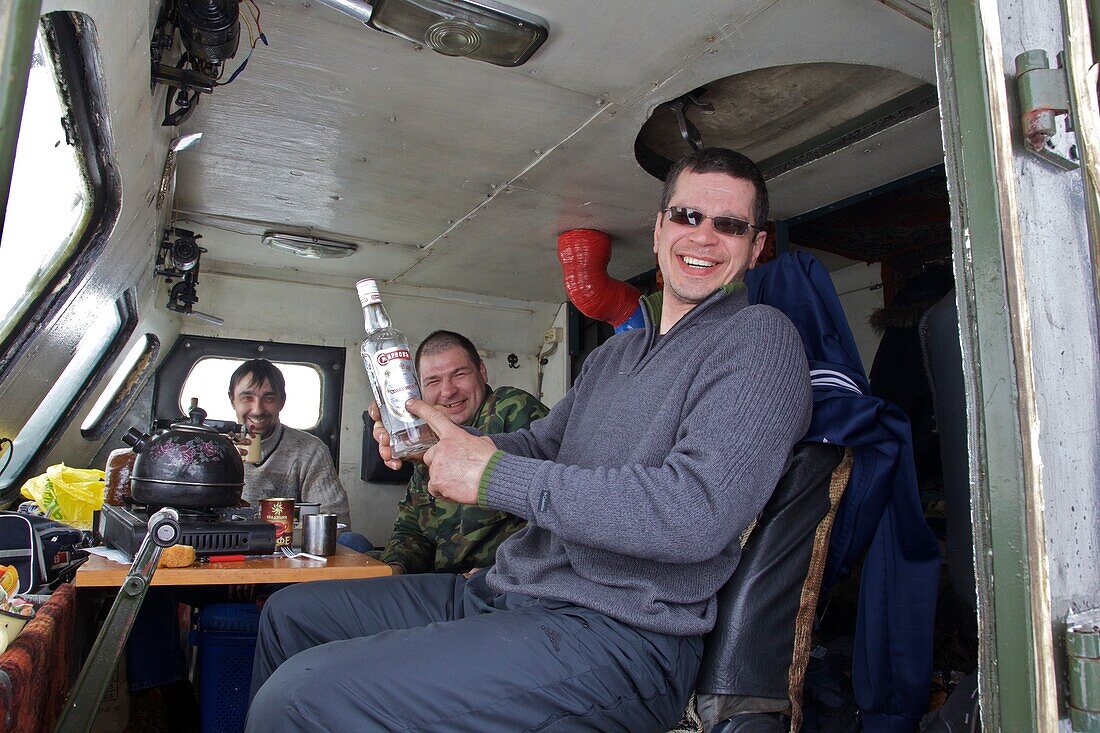 three men in the operator's cab of a tracked vehicle, one laughing at a bottle of vodka, Chukotka Autonomous Okrug, Siberia, Russia