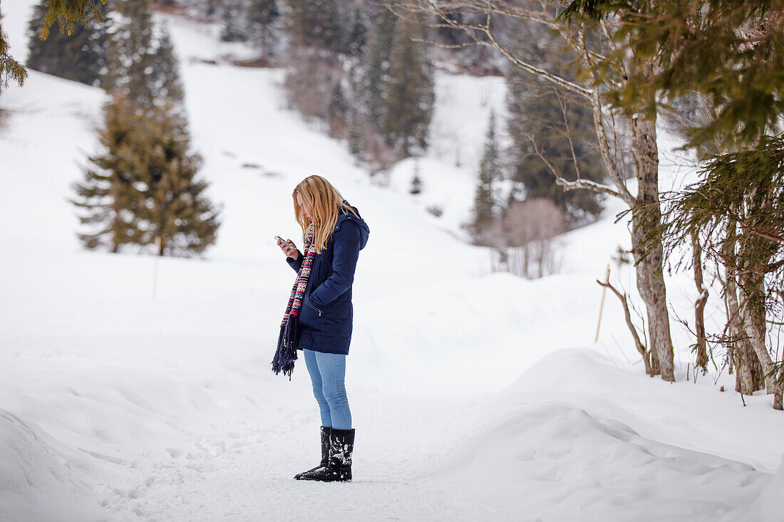 Junge Frau mit einem Handy steht im Schnee, Spitzingsee, Oberbayern, Bayern, Deutschland