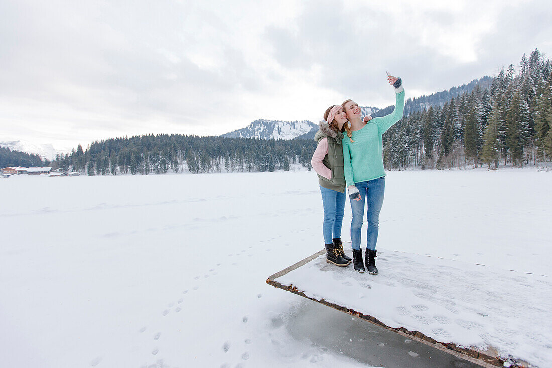 Two young women on jetty photographing themselves, Spitzingsee, Upper Bavaria, Germany