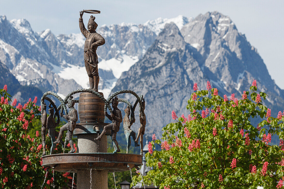 fountain with sculpture of traditional Coopers Dance, Wetterstein mountains, Zugspitze, Chestnut blossom in Spring, Partenkirchen, Garmisch-Partenkirchen, Werdenfelser Land, Baverian Alps, Upper Baveria, Bavaria, Germany, Europe