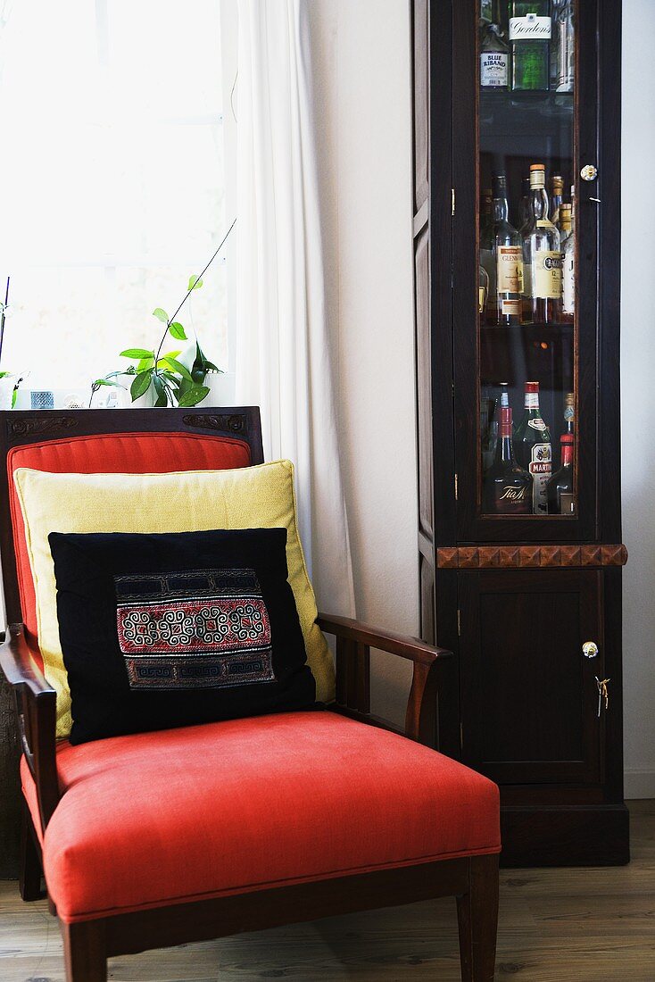 A red upholstered armchair with cushions and bottles of alcohol in a cabinet