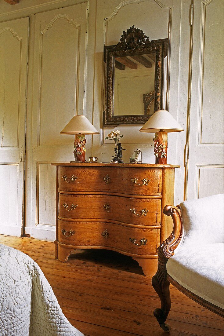 Table lamps on a Rococo chest of drawers with a mirror hanging above it in front of a wood panelled wall