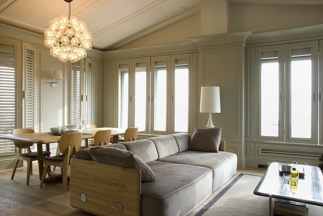 Sofa and dining area with hanging lamps in front of a wall of floor to ceiling windows and bright gray wood paneling