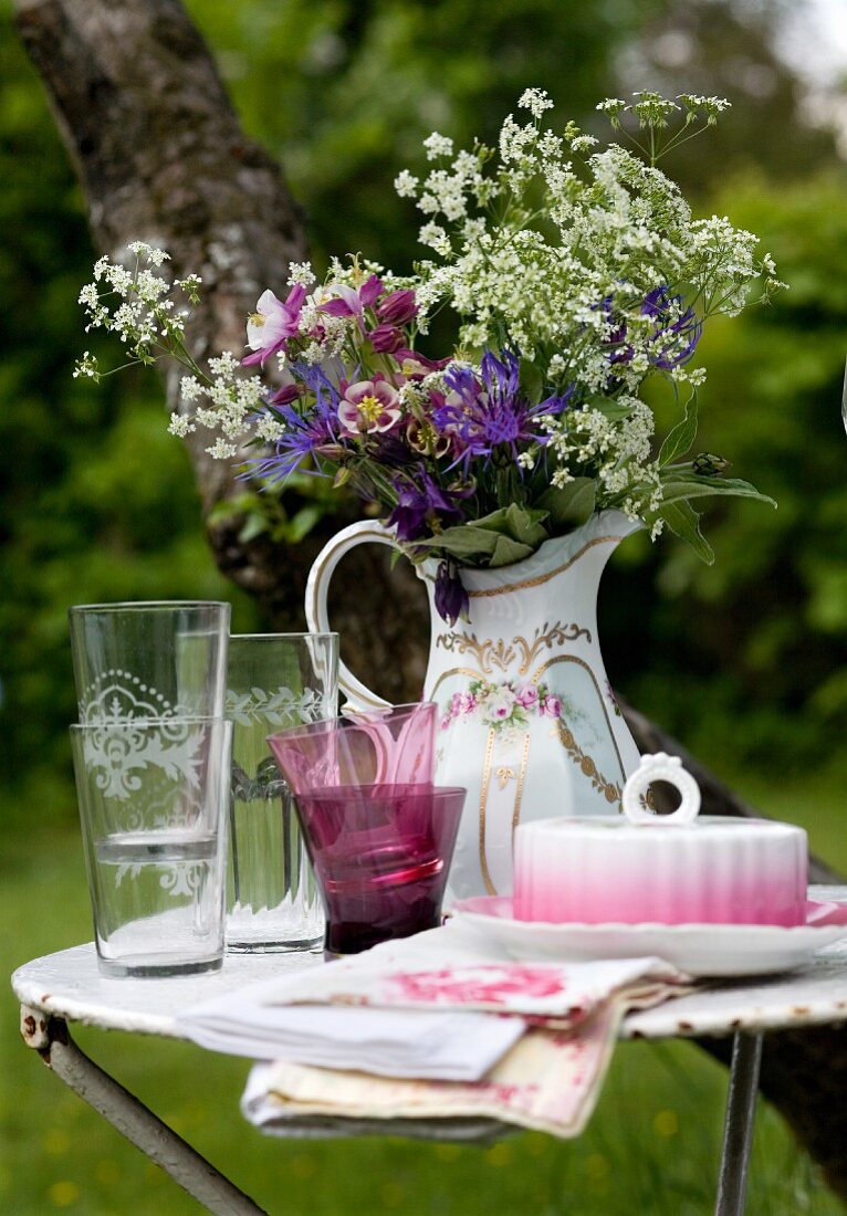 Glasses and butter dish in front of a pitcher filled with garden flowers