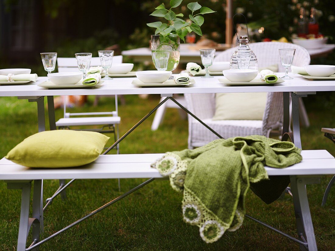 A table laid in a garden with a white bench and green cushions and a blanket