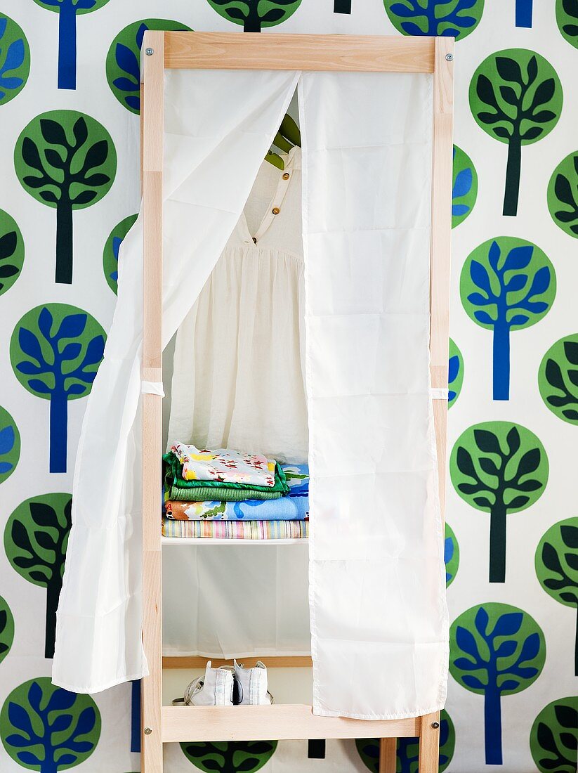 A wooden shelf against a wall with floral patterned wall paper with an open white curtain and a view of clothes