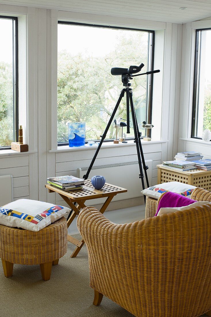 Rattan chair and foot stool around a wooden side table next to a tripod with camera in front of a bank of windows