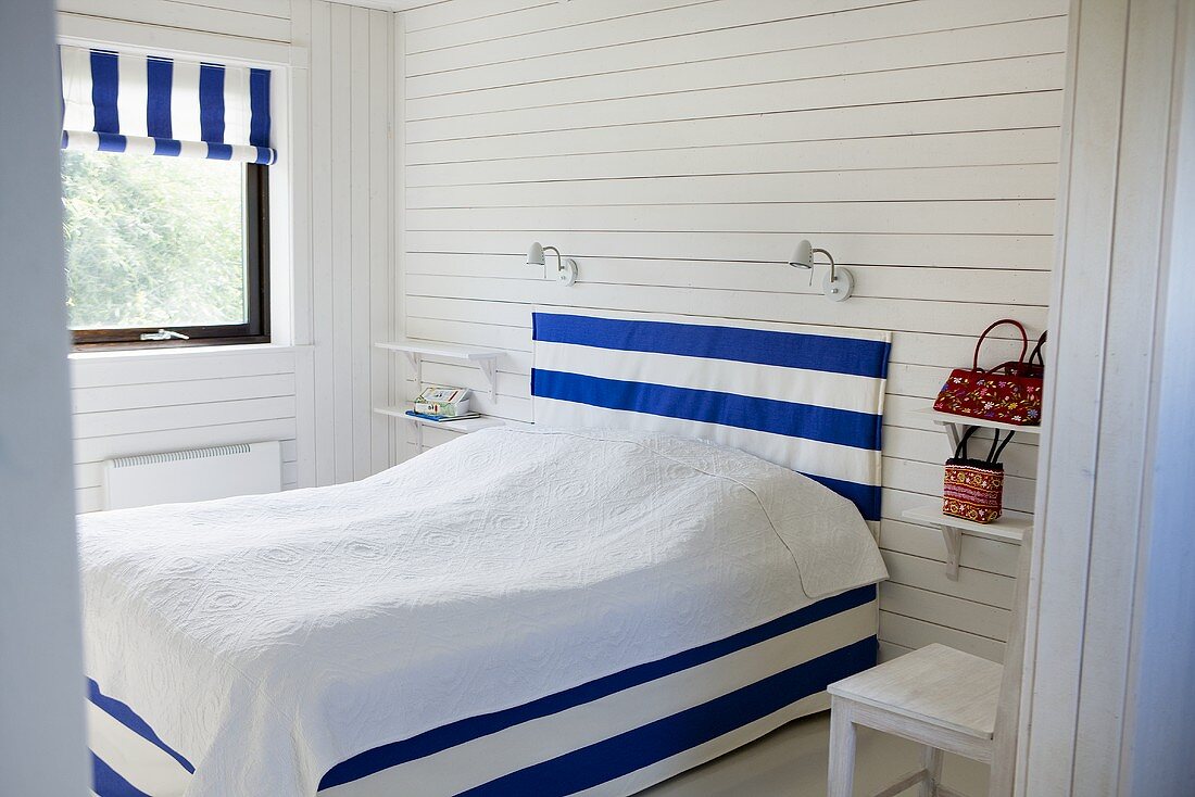 Bedroom with white wood paneling and double bed with cover, head board and blind with a blue and white design