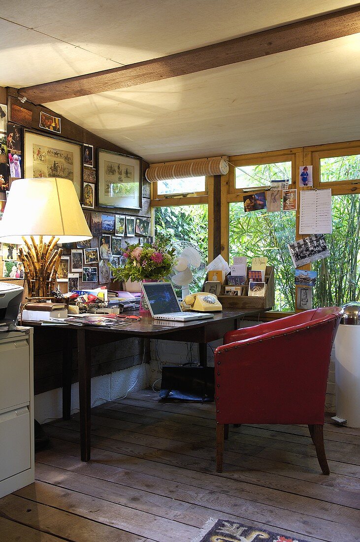 A home office space inside a shed with a wooden desk and a red chair upholstered chair on wooden floorboards.