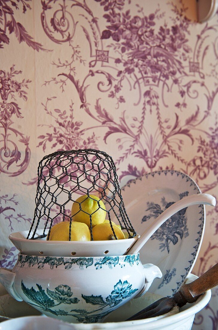 A wire basket over lemons on a ceramic sieve and a stack of plates against a floral-patterned wallpaper