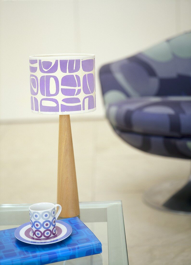 A detail of a modern, retro styled sitting room, showing a wood lamp with lilac pattern shade, glass table, cup and saucer