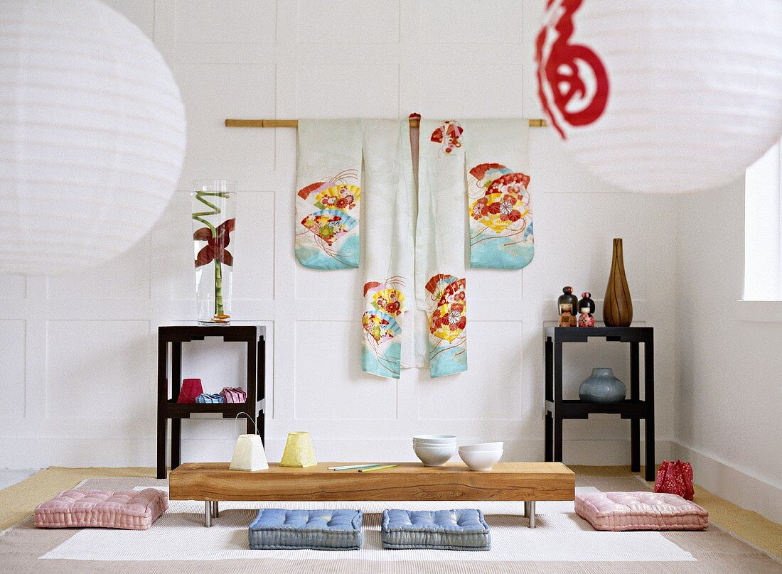 Japanese sitting room with a white panelled wall, hanging kimono, paper lanterns, black lacquered side tables, and a low wooden table with floor cushions.