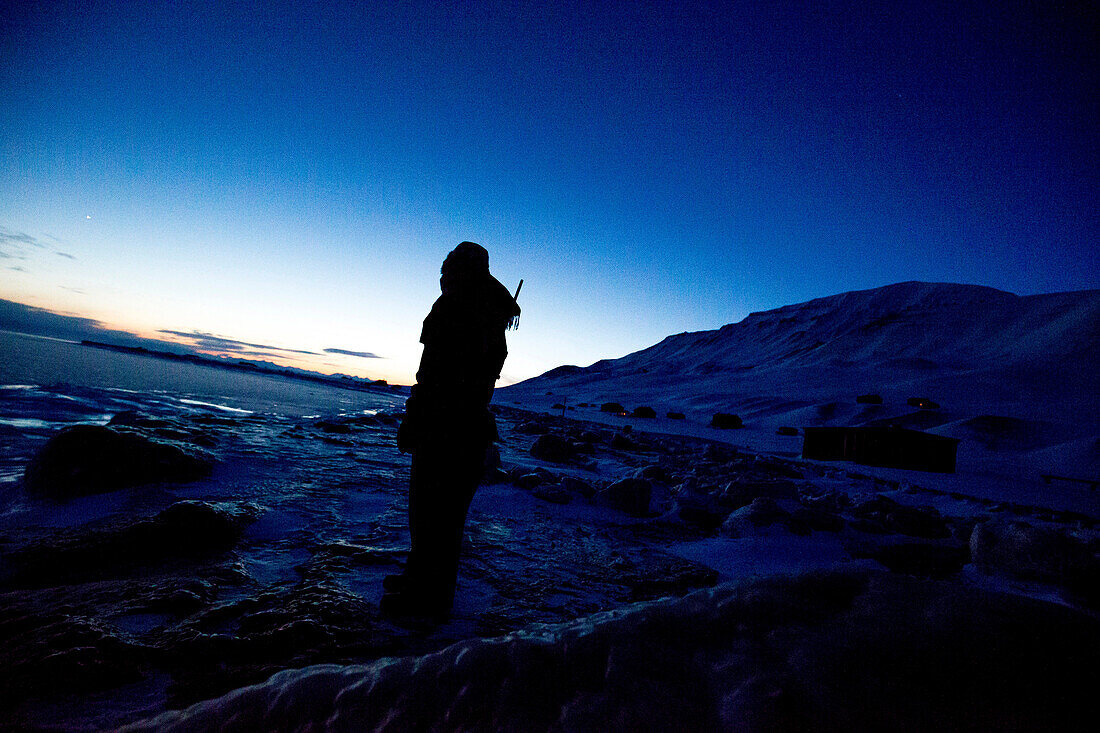 Person mit Waffe am Rücken schaut in die winterliche Landschaft bei Nacht, Spitzbergen, Svalbard, Norwegen