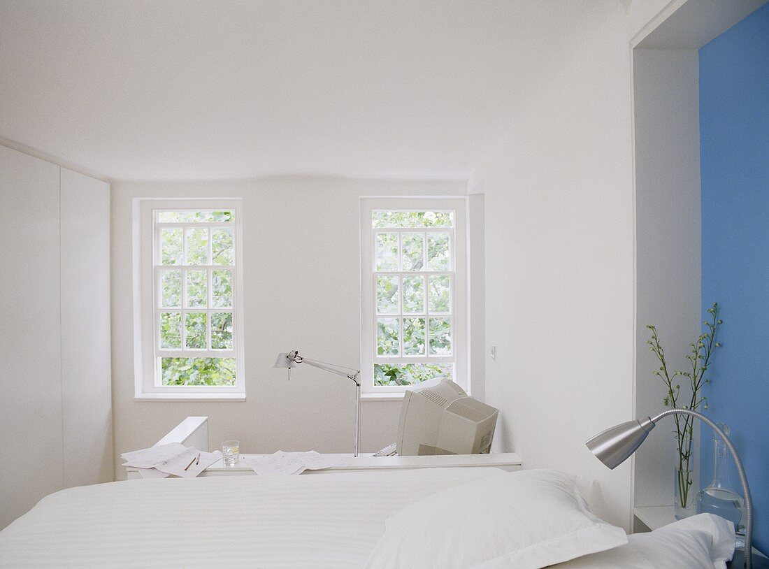 A modern, white open bedroom with office area, blue wall, double bed, desk with computer, plain window,