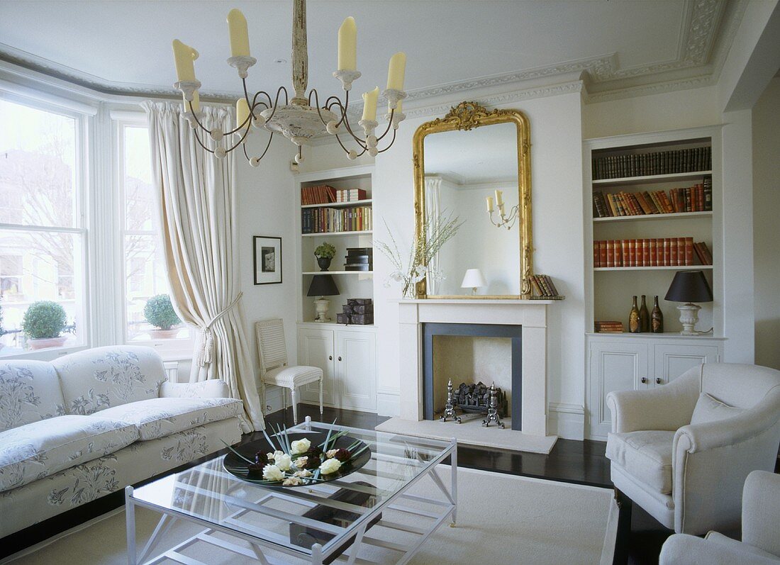 A traditional, neutral sitting room, with fireplace, gilt mirror, book shelves and modern glass coffee table
