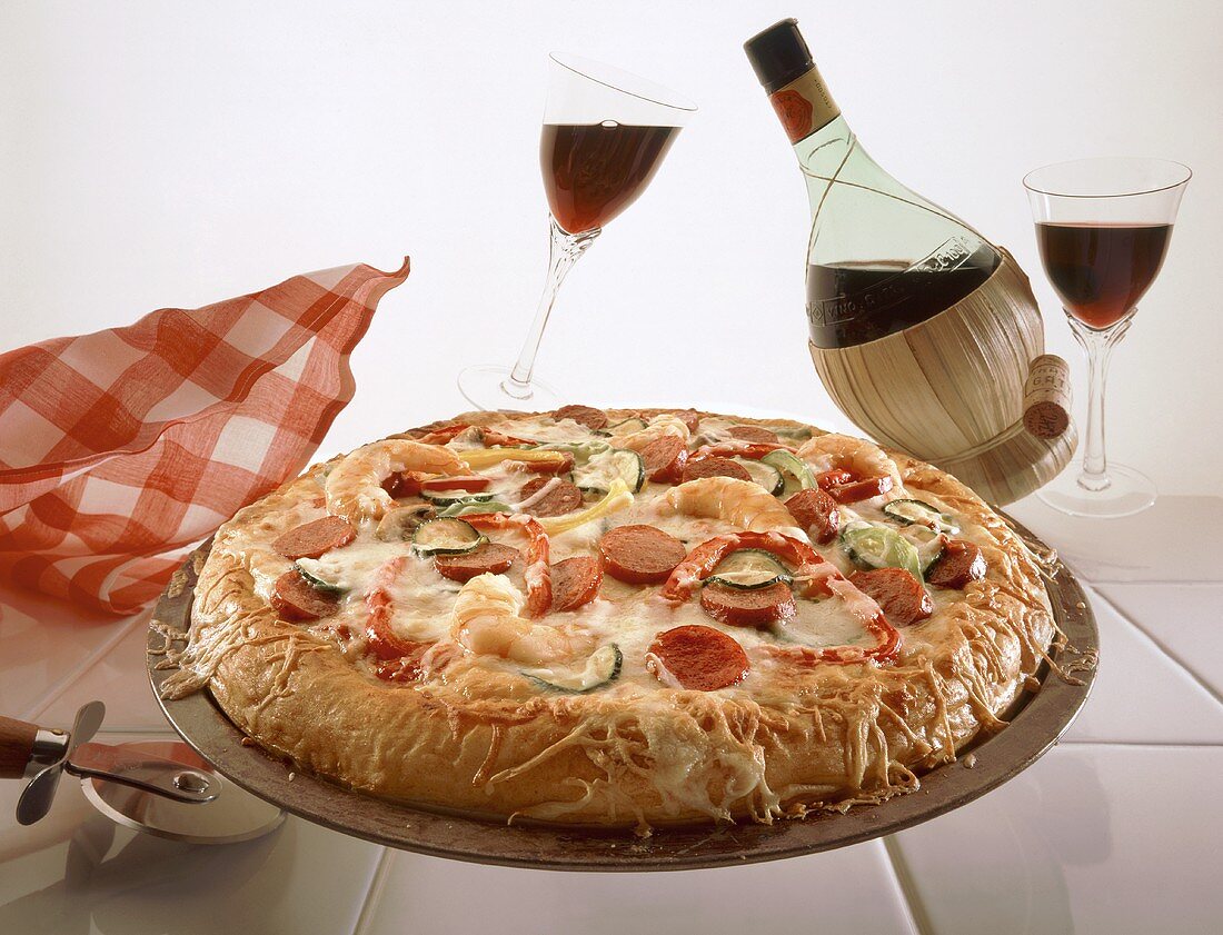 Pizza mit Schrimps, Wurst und Gemüse, dahinter Rotwein