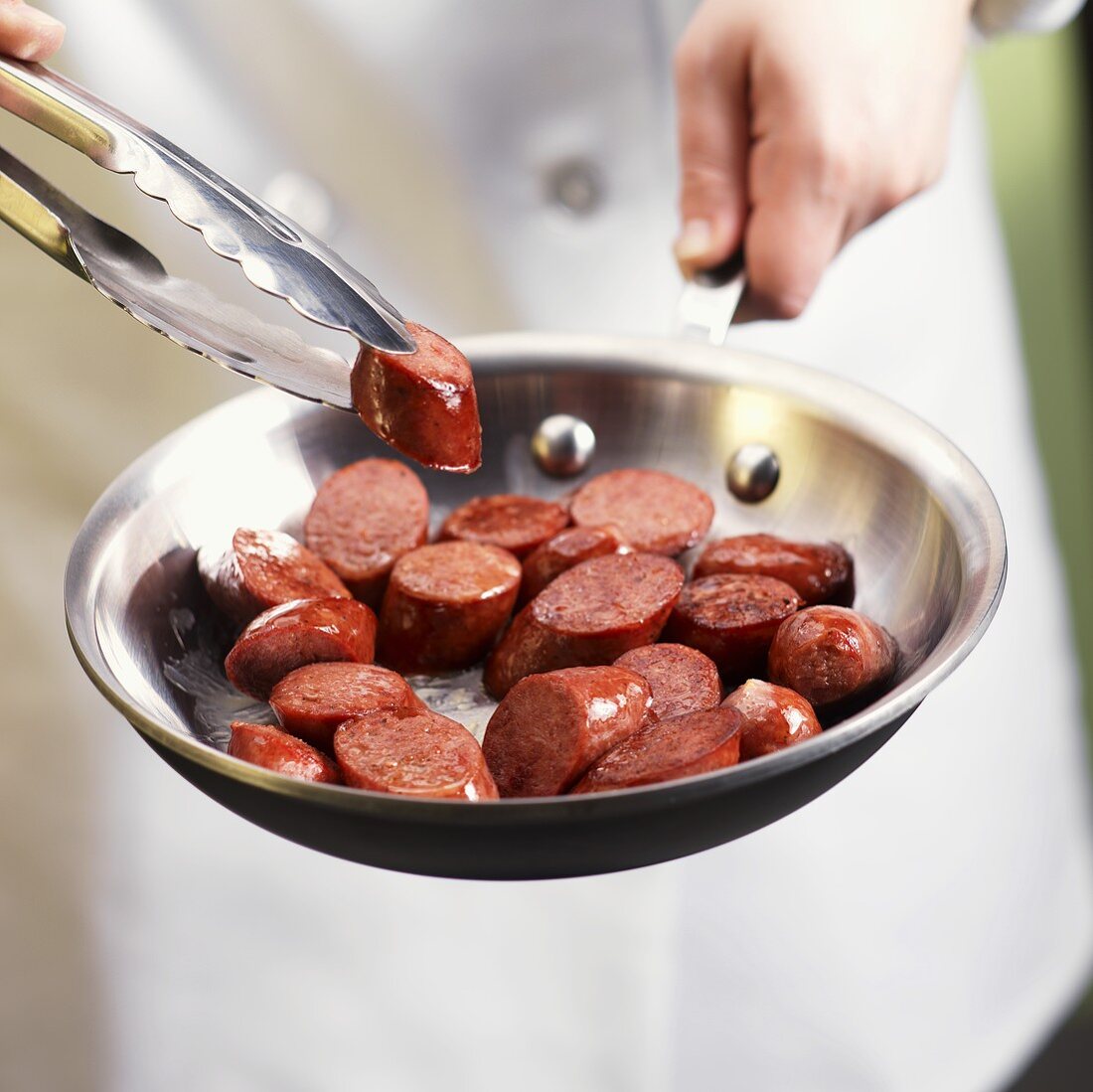 Hands Holding Tongs with a Piece of Andouille Sausage over Skillet