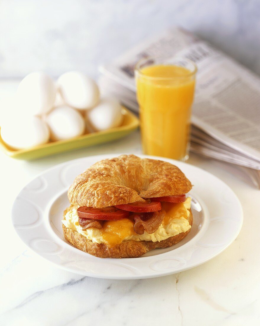 Croissant mit Rührei, Bacon und Tomaten, Orangensaft, Eier, Zeitung